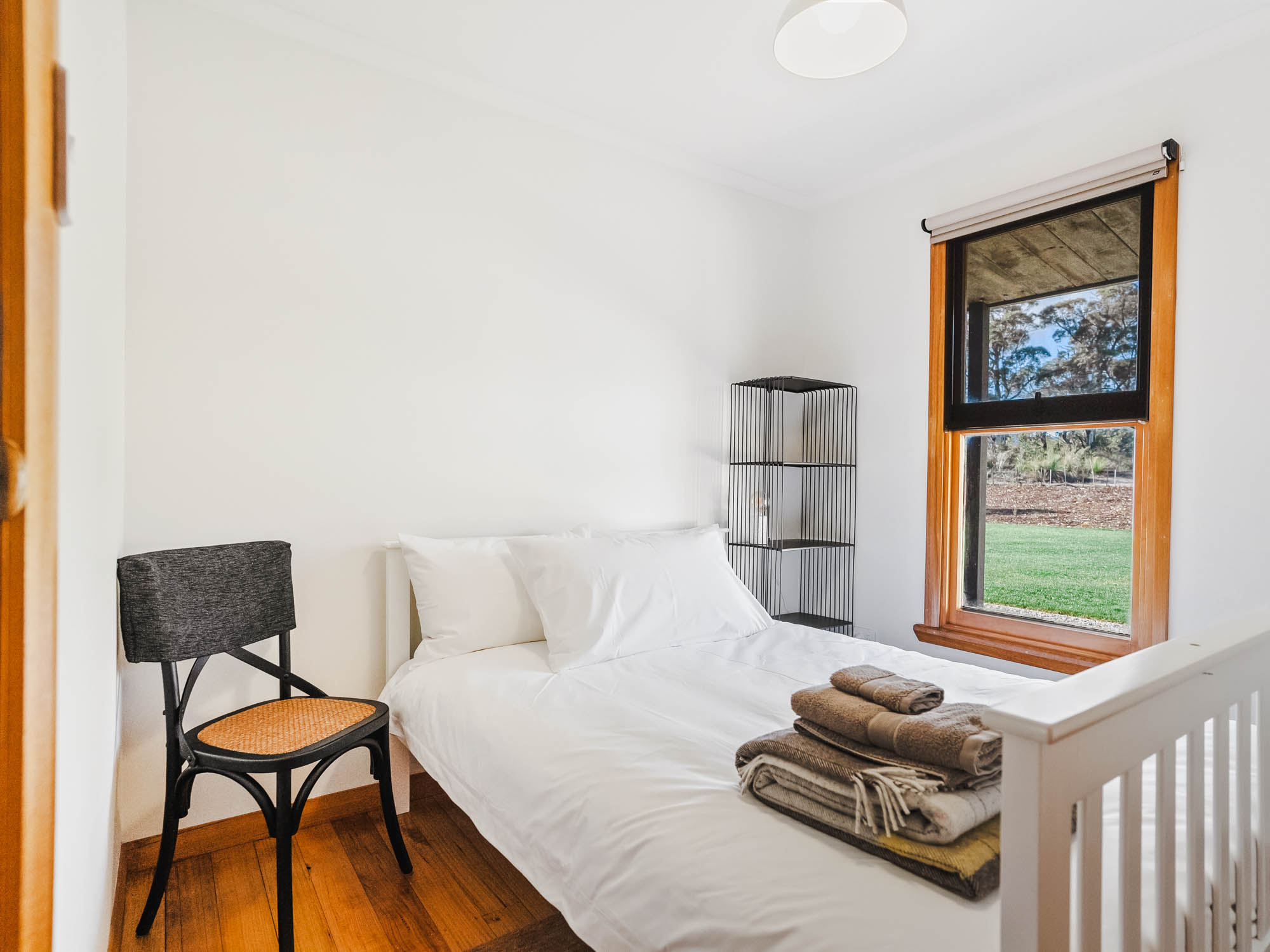 Small bedroom with white bed and pillows, folded towels on the bed, a black chair with a woven seat, and a window showing green grass outside.