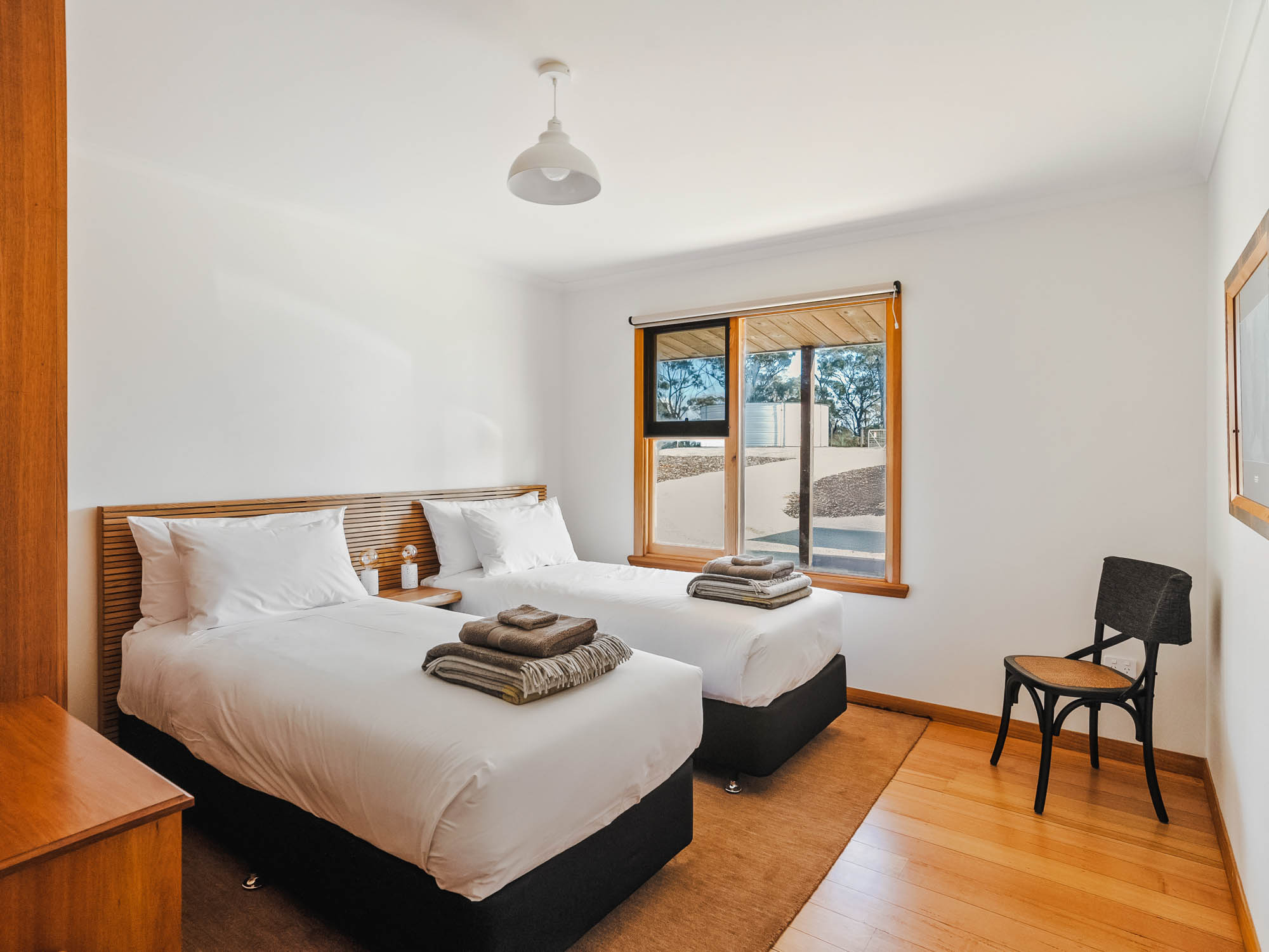 Bedroom with two single beds with white linens, a wooden headboard, folded towels on each bed, a window with a view outside, a black chair, and wooden flooring.