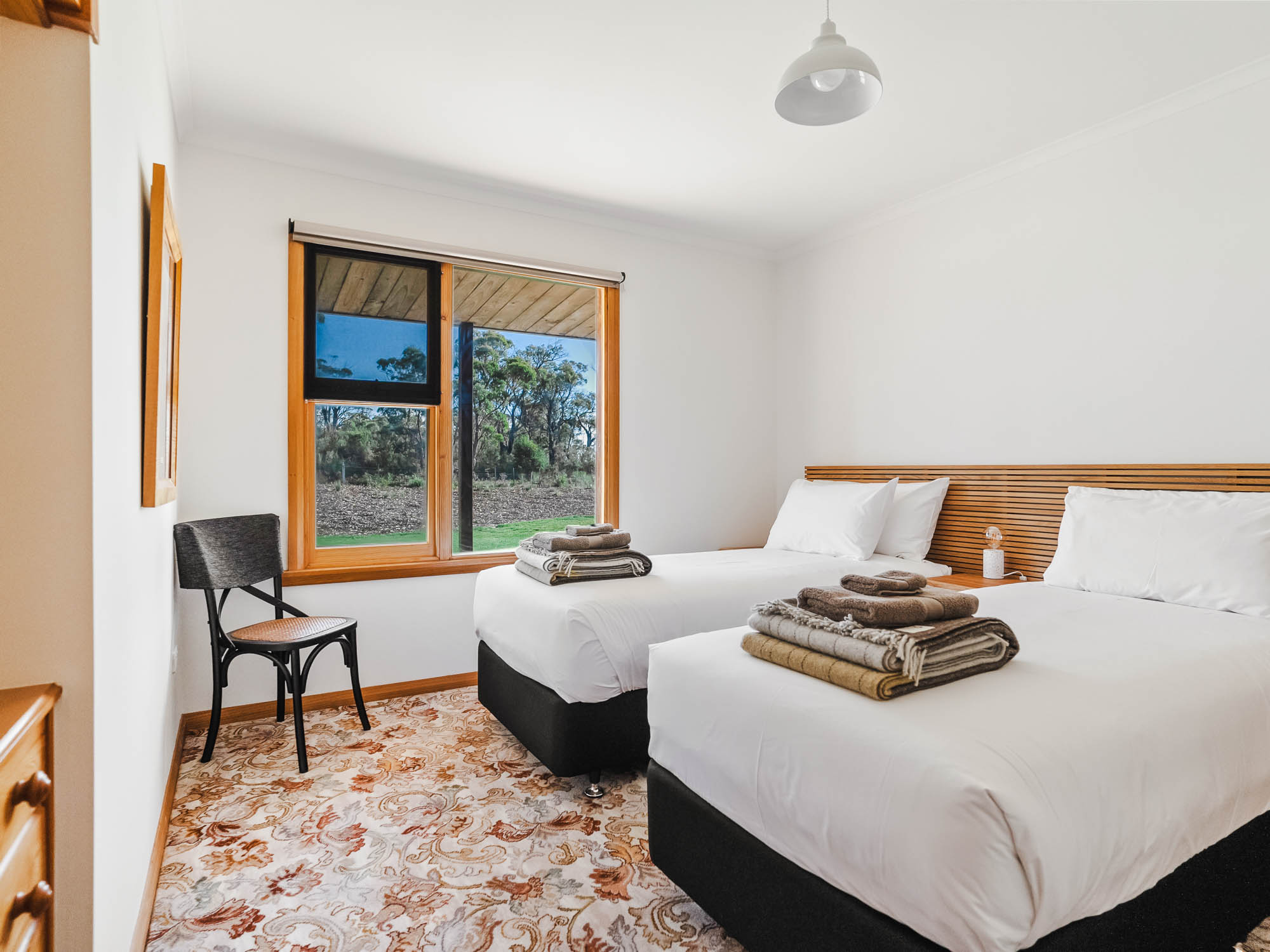 Bedroom with two single beds, patterned carpet, a window showing trees outside, and a black chair.