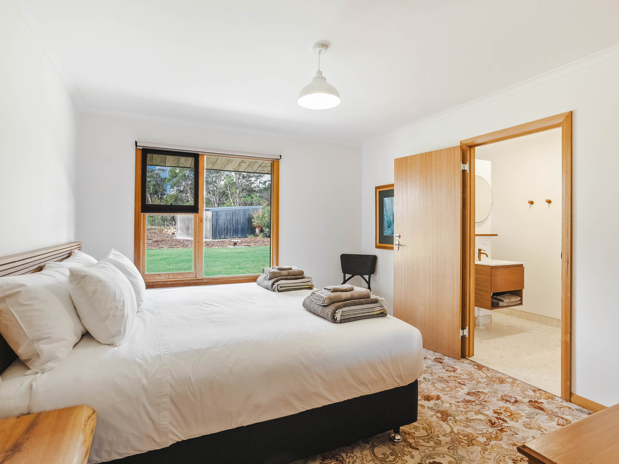Bright bedroom with white walls, a large bed with white linens, folded towels on the bed, a window showing a green lawn outside, and an open door leading to a bathroom.