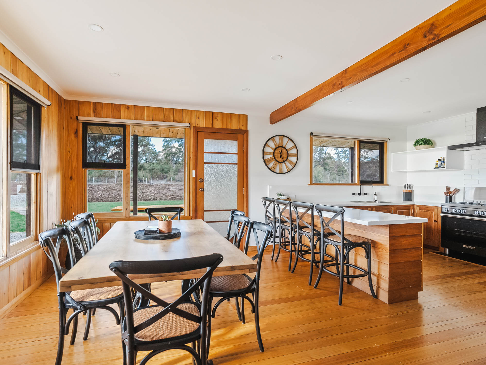 Bright kitchen and dining area with wooden floors and walls, a wooden dining table with black chairs, a kitchen island with stools, and large windows showing outdoor greenery.