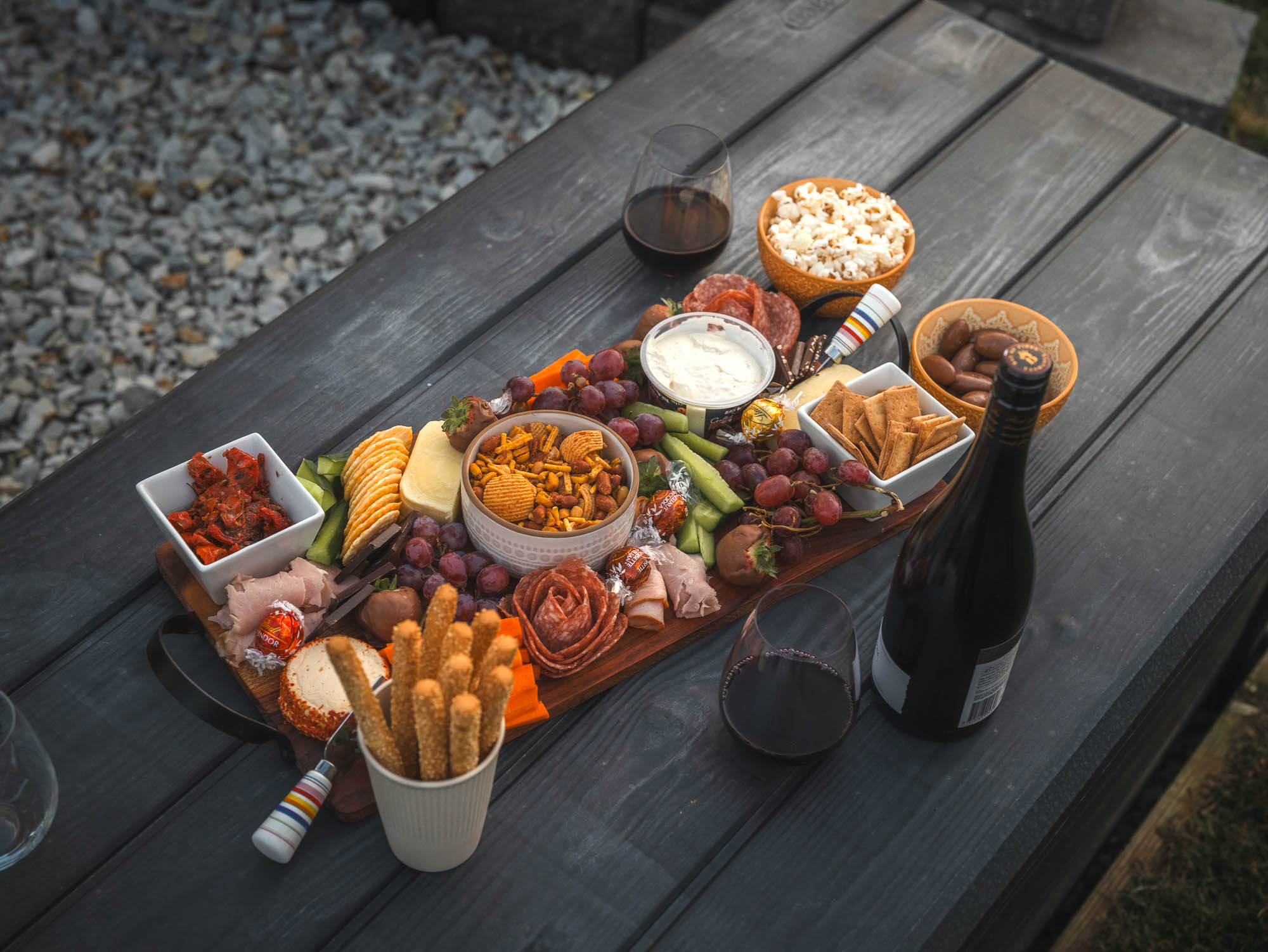 Charcuterie board with assorted cheeses, meats, grapes, crackers, dips, and snacks on a wooden table with glasses and a bottle of red wine.