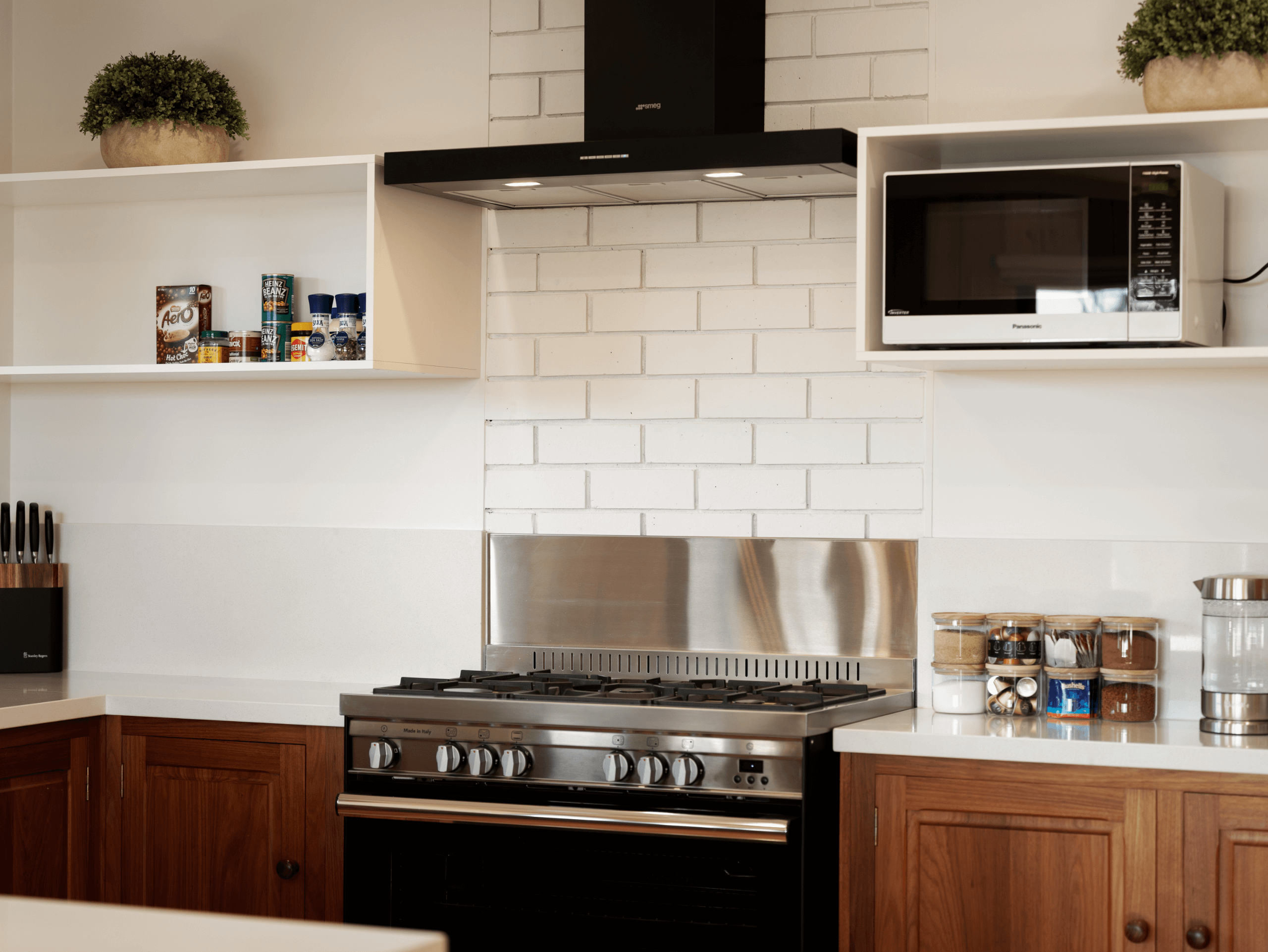Modern kitchen with gas stove, black range hood, microwave, wooden cabinets, spice jars, and potted plants on shelves.
