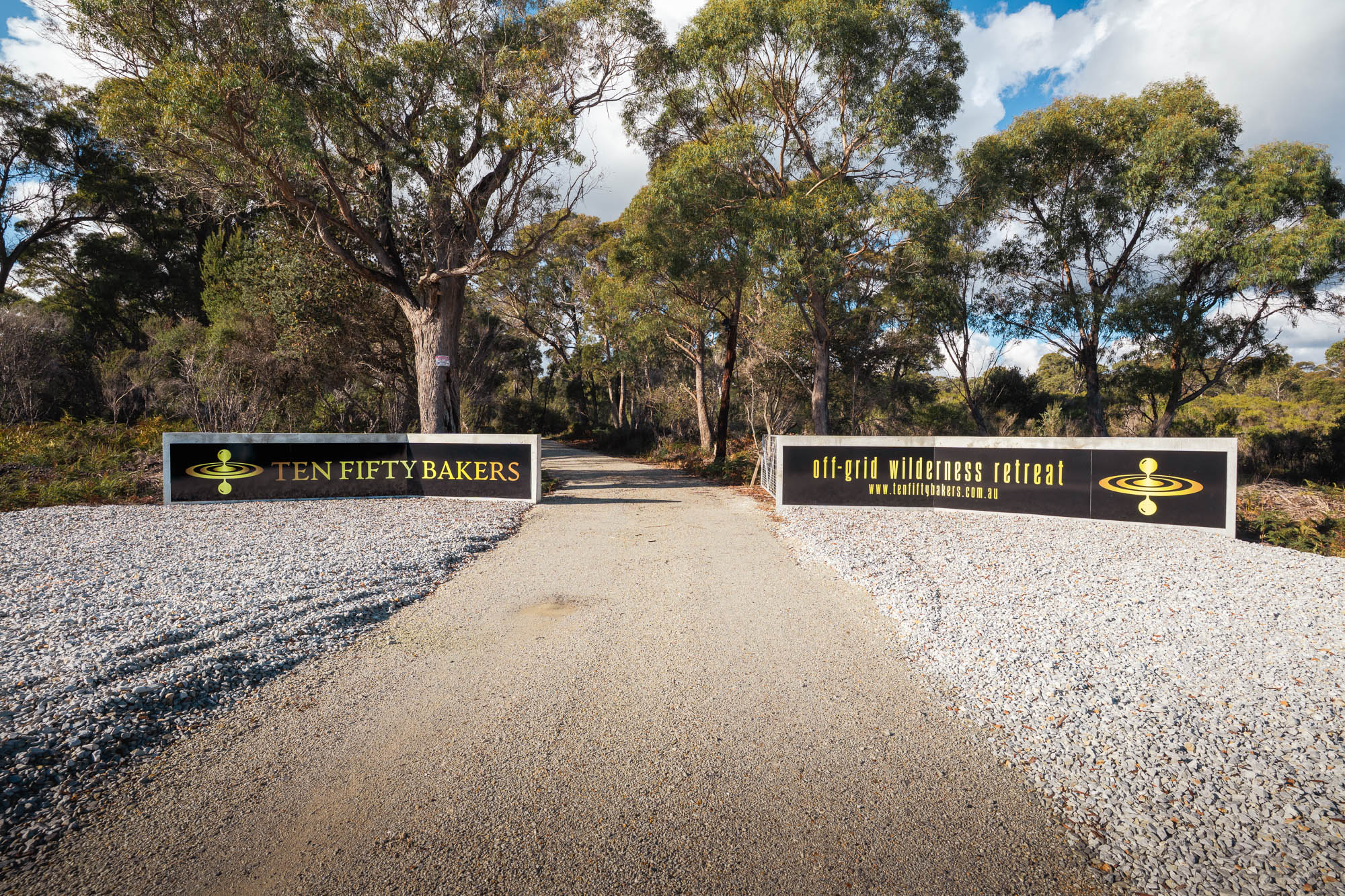 Gravel driveway entrance flanked by signs for Ten Fifty Bakers off-grid wilderness retreat surrounded by trees under a partly cloudy sky.