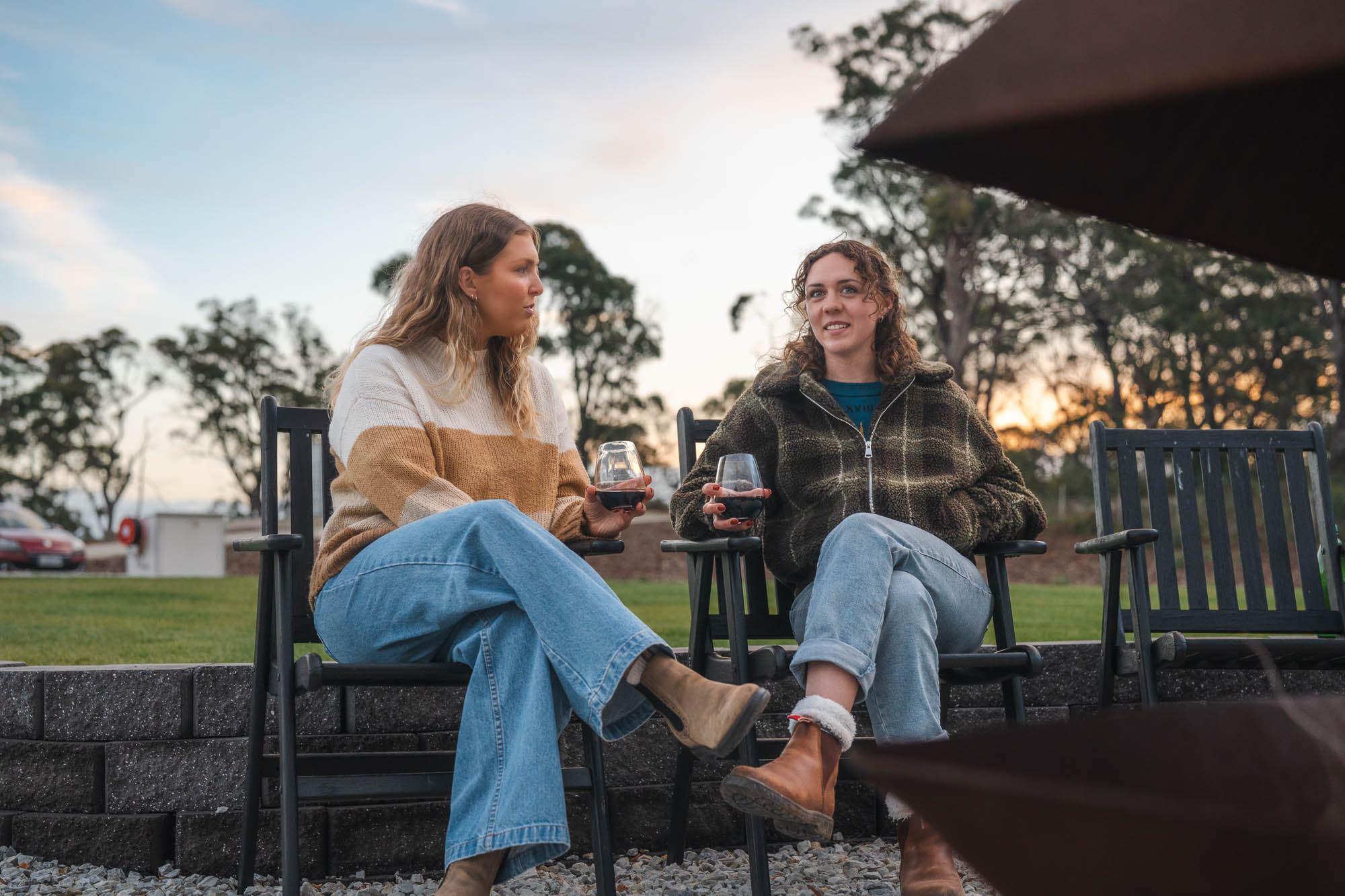 Two women sitting outside on black chairs, drinking red wine and talking at sunset.