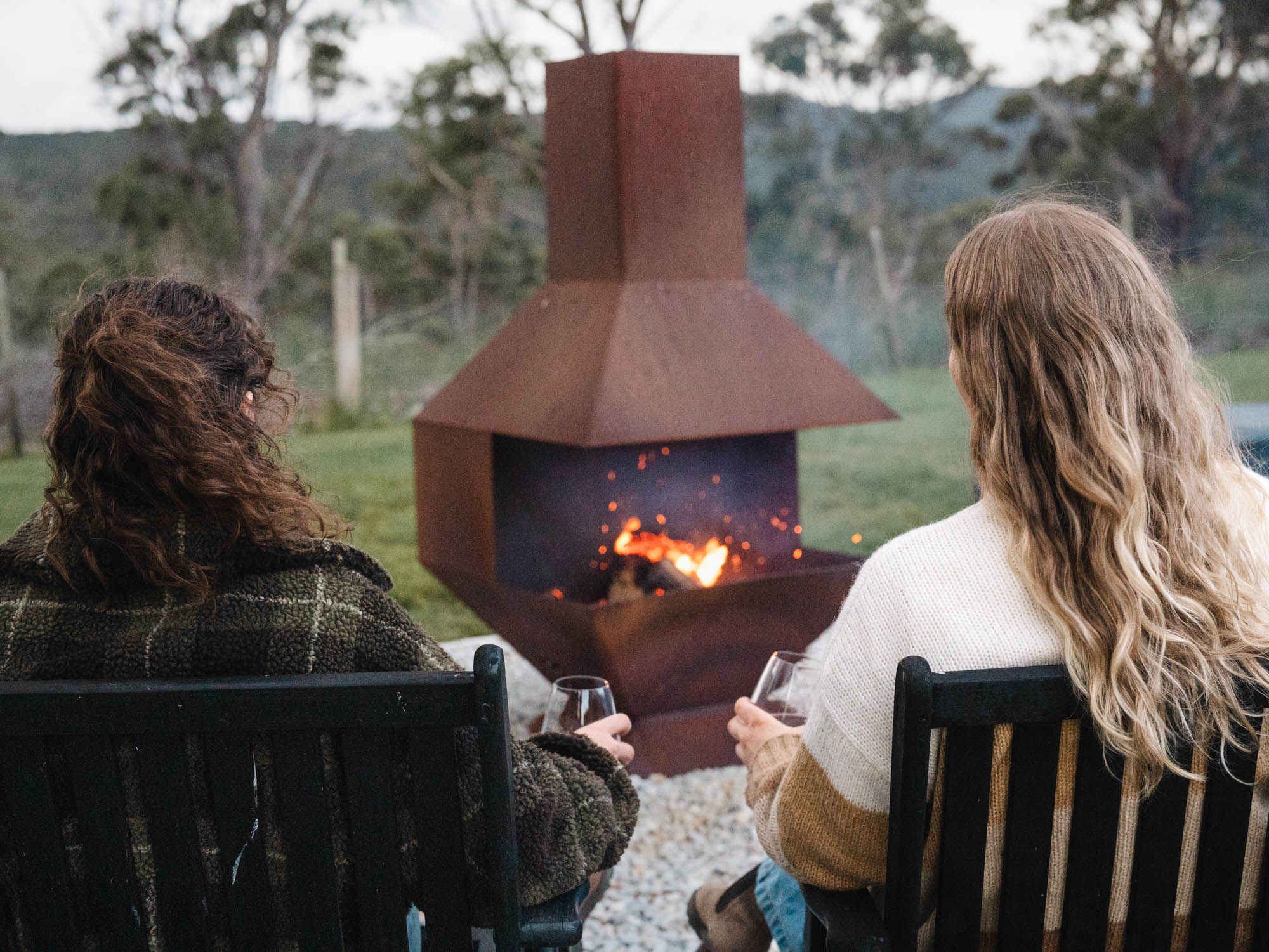 Two people sitting on chairs holding wine glasses in front of an outdoor fire pit with a fire burning.
