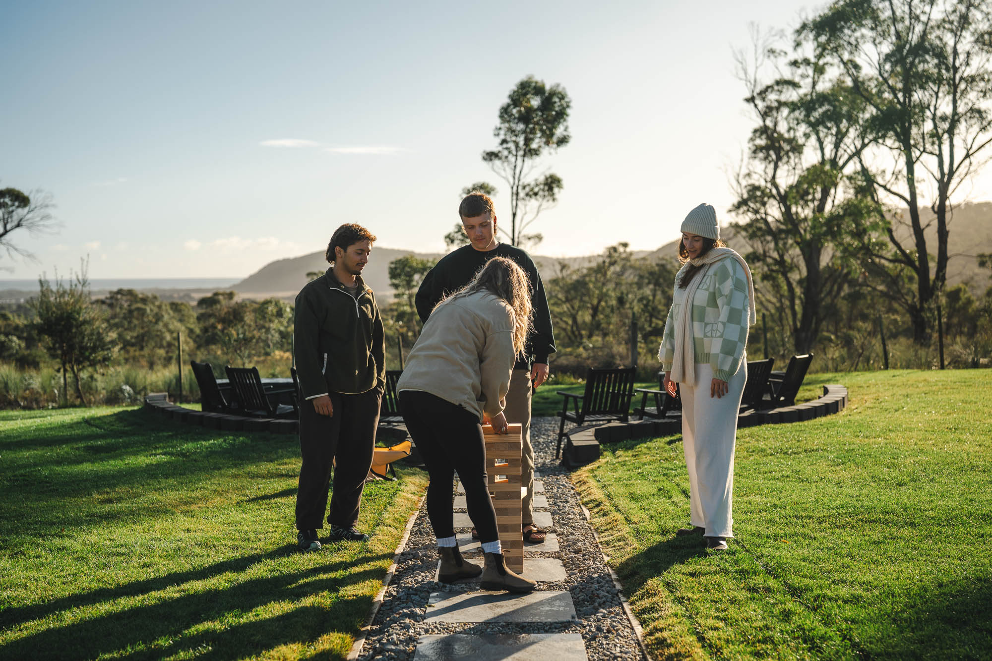 Four people playing giant Jenga outdoors on a stone path with trees and hills in the background during daylight.