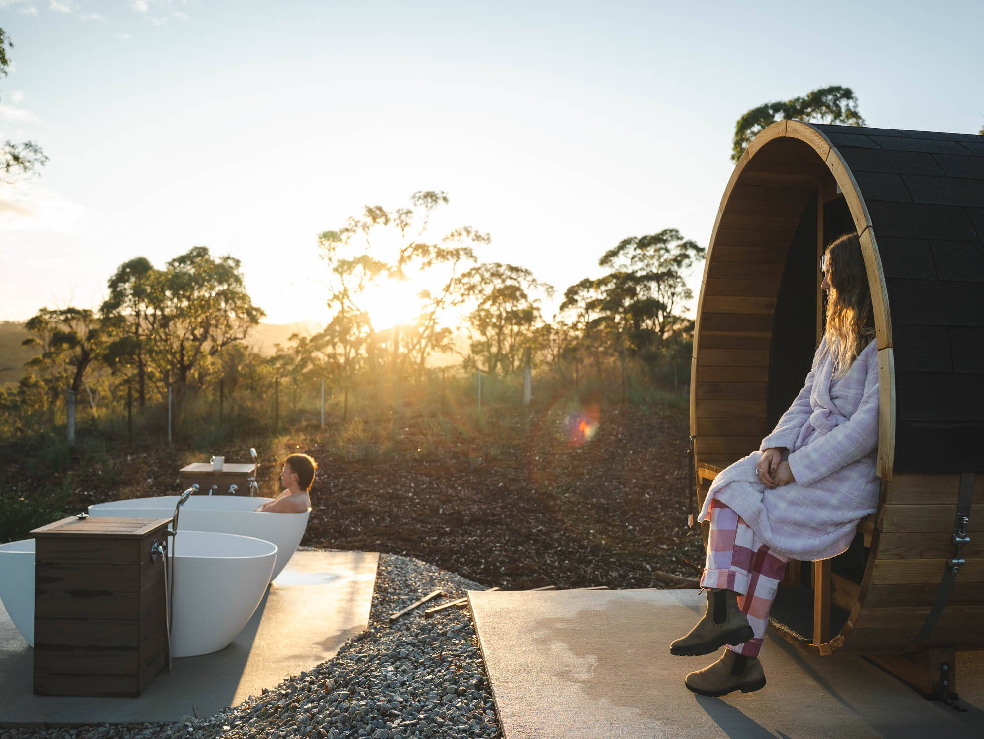 A person relaxing in an outdoor white bathtub at sunset while another person in a robe sits on a wooden barrel sauna nearby surrounded by trees.