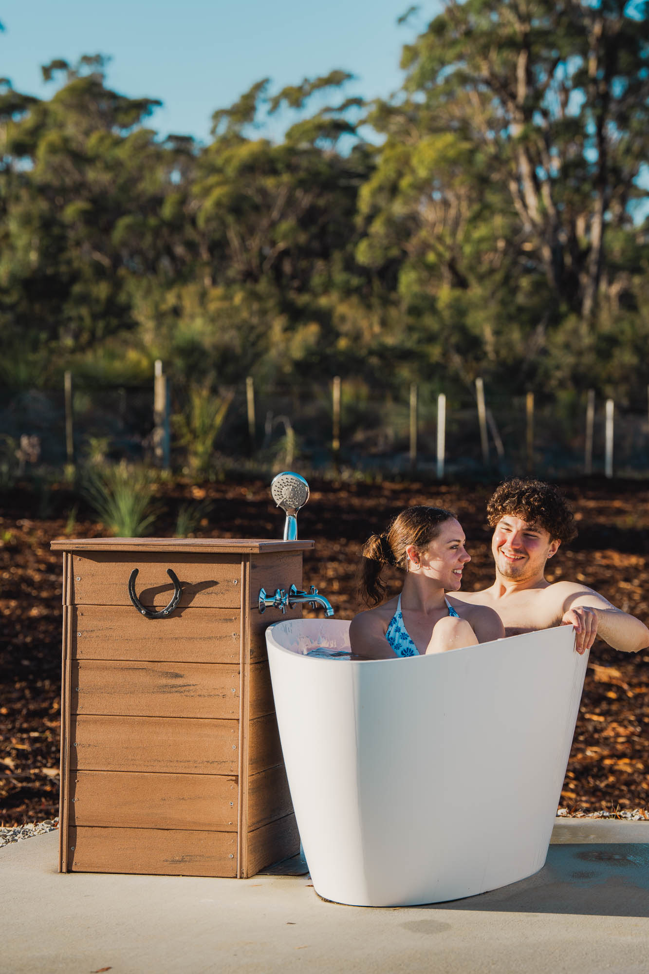 Young couple smiling and sitting together in an outdoor white bathtub near a wooden shower structure.
