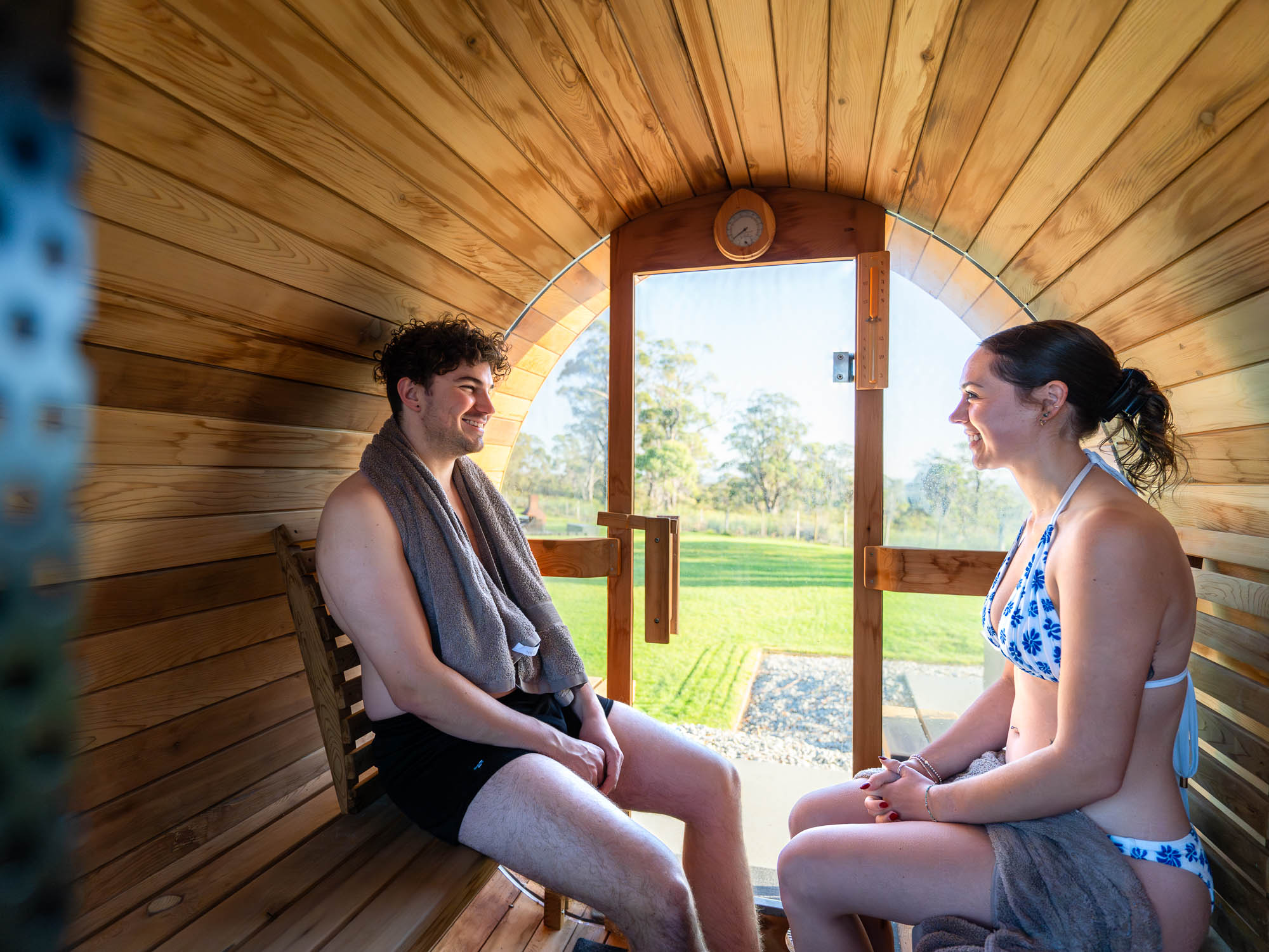 Man and woman sitting inside a wooden barrel sauna, smiling and facing each other with a glass door showing a green outdoor landscape.
