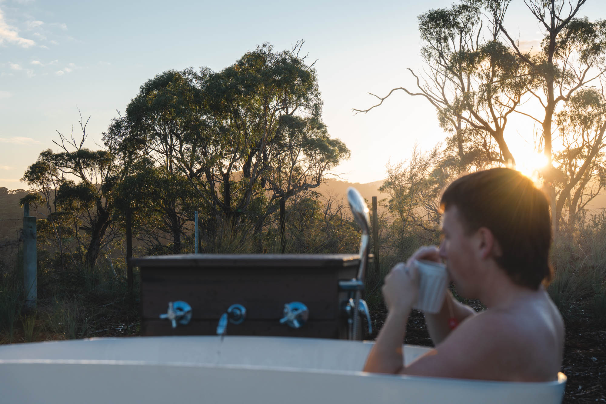Person drinking from a mug while sitting in an outdoor bathtub at sunset with trees in the background.