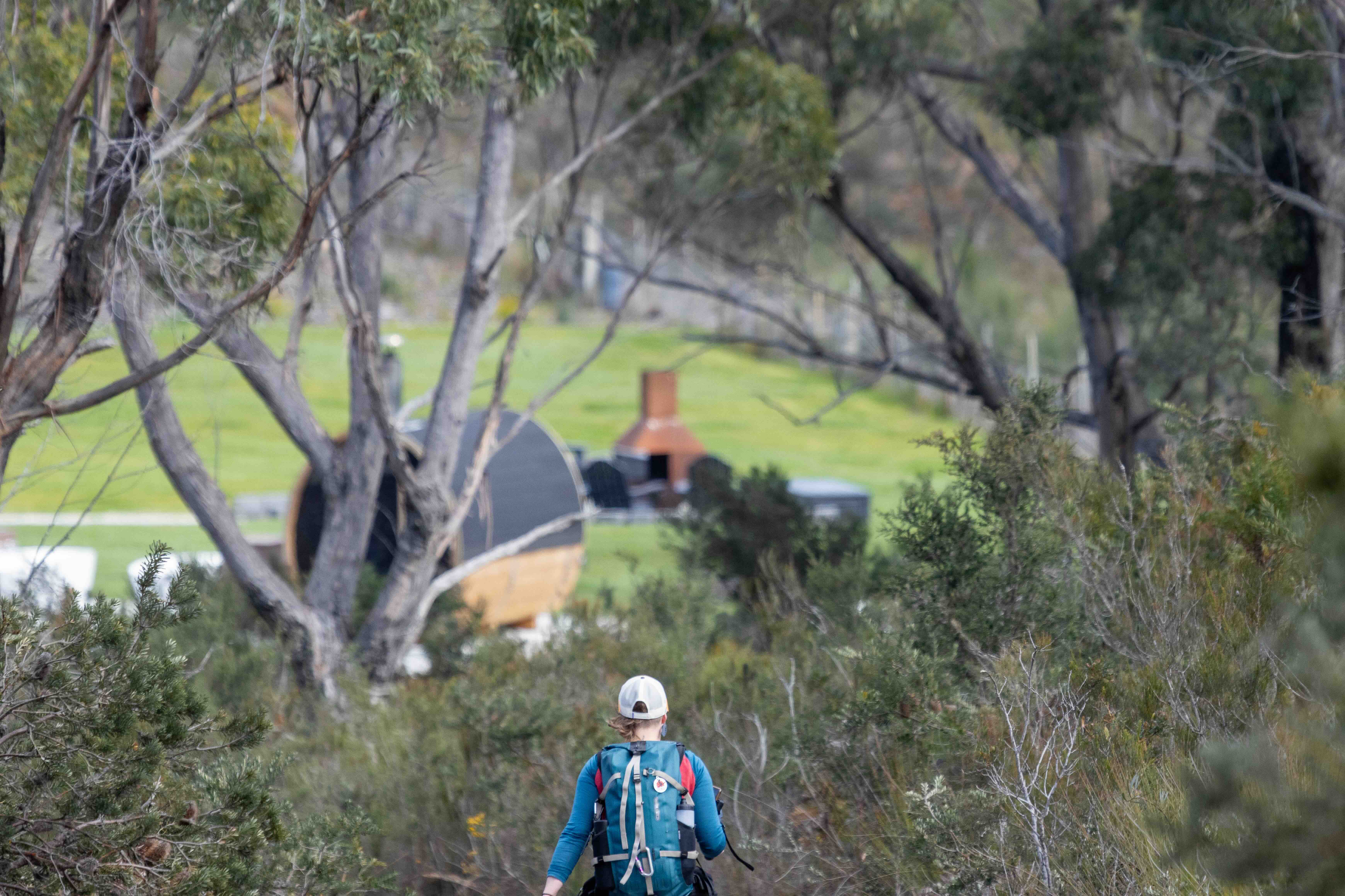 Person wearing a blue jacket and white cap hiking on a trail surrounded by trees and bushes, heading towards a grassy area with outdoor furniture and a chimney.