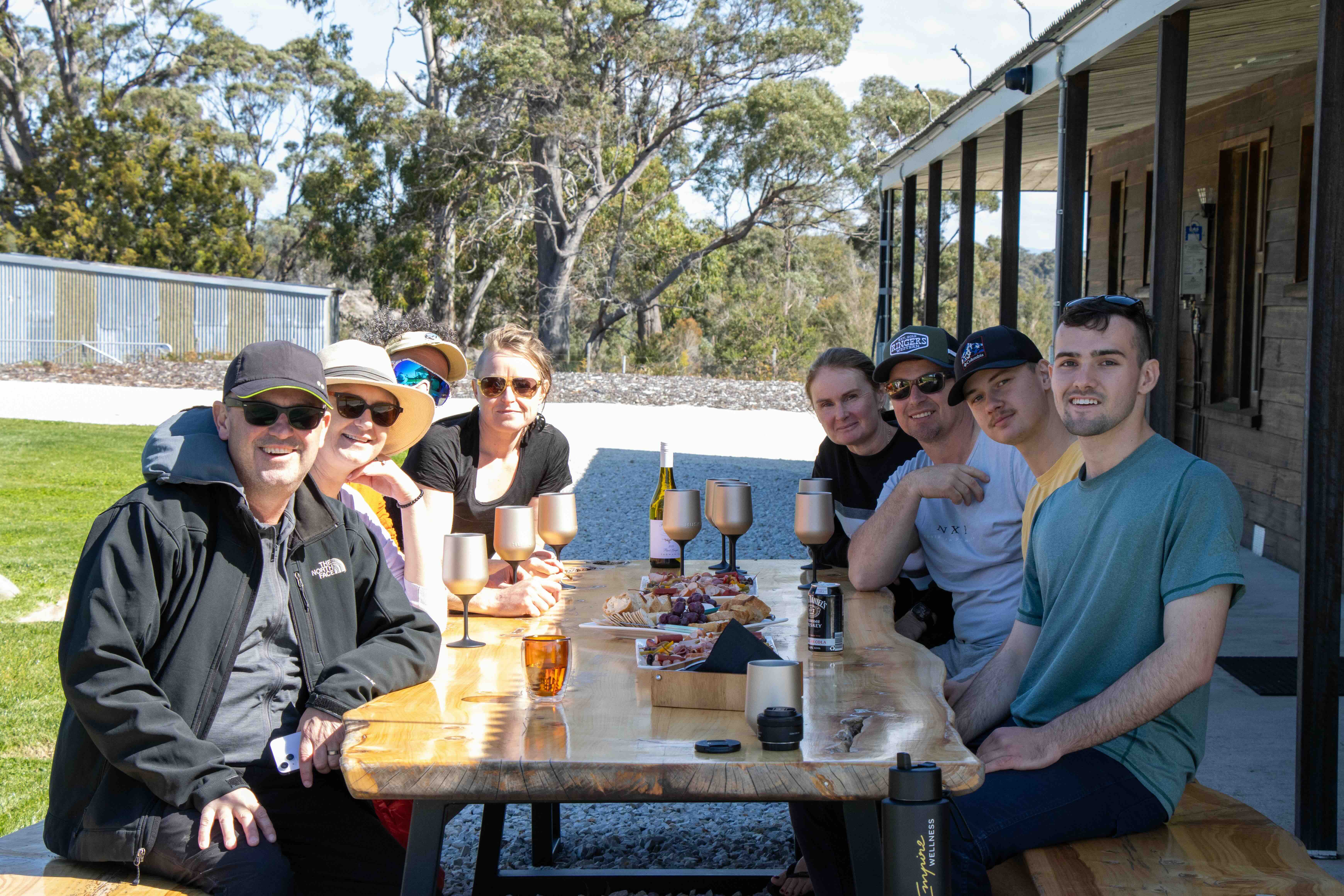 Group of eight people sitting at a wooden table outdoors with wine glasses and platters of snacks, smiling at the camera.