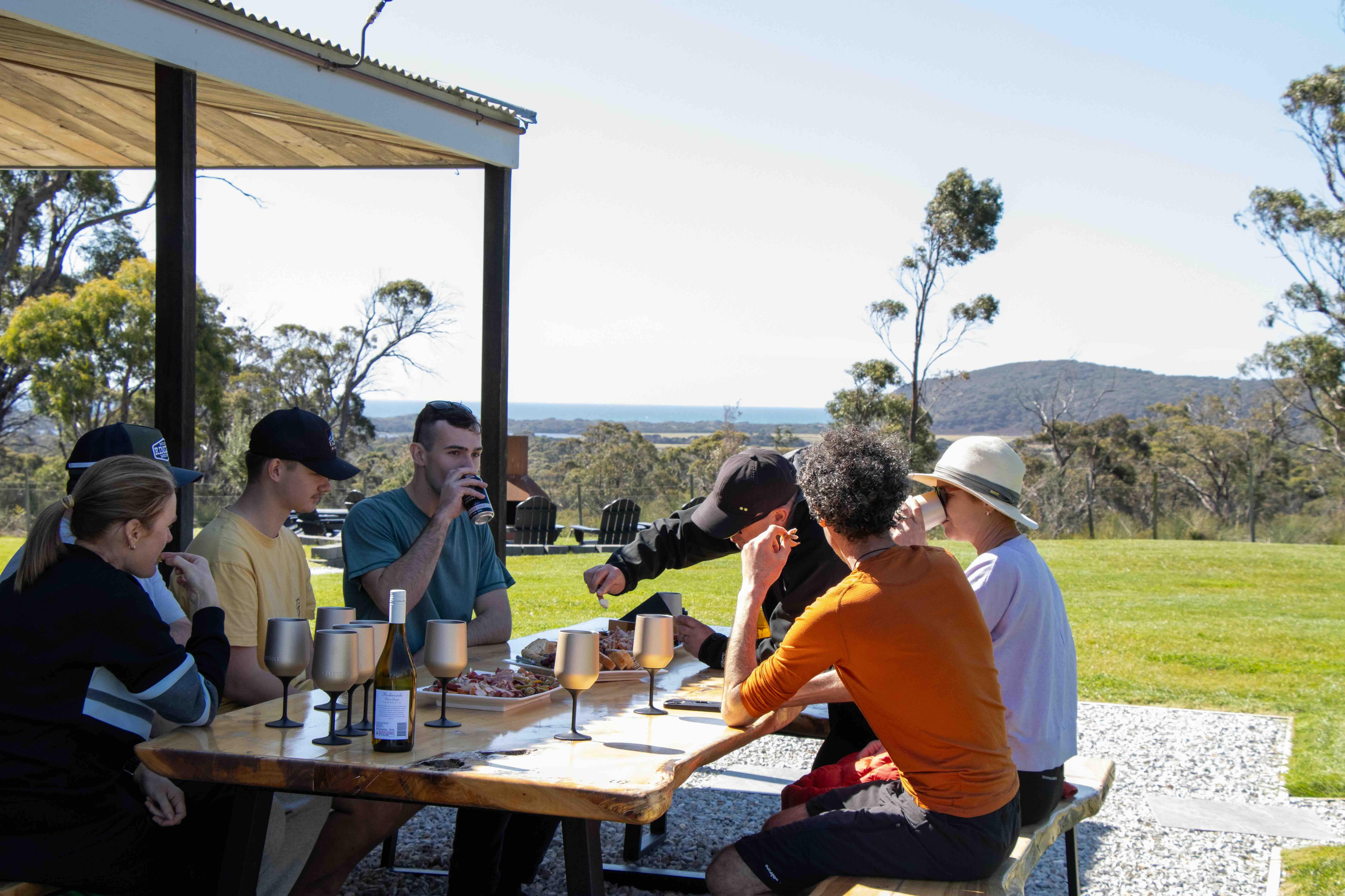Group of six people sitting outdoors at a wooden table with drinks and snacks, overlooking a grassy landscape with trees and distant hills.