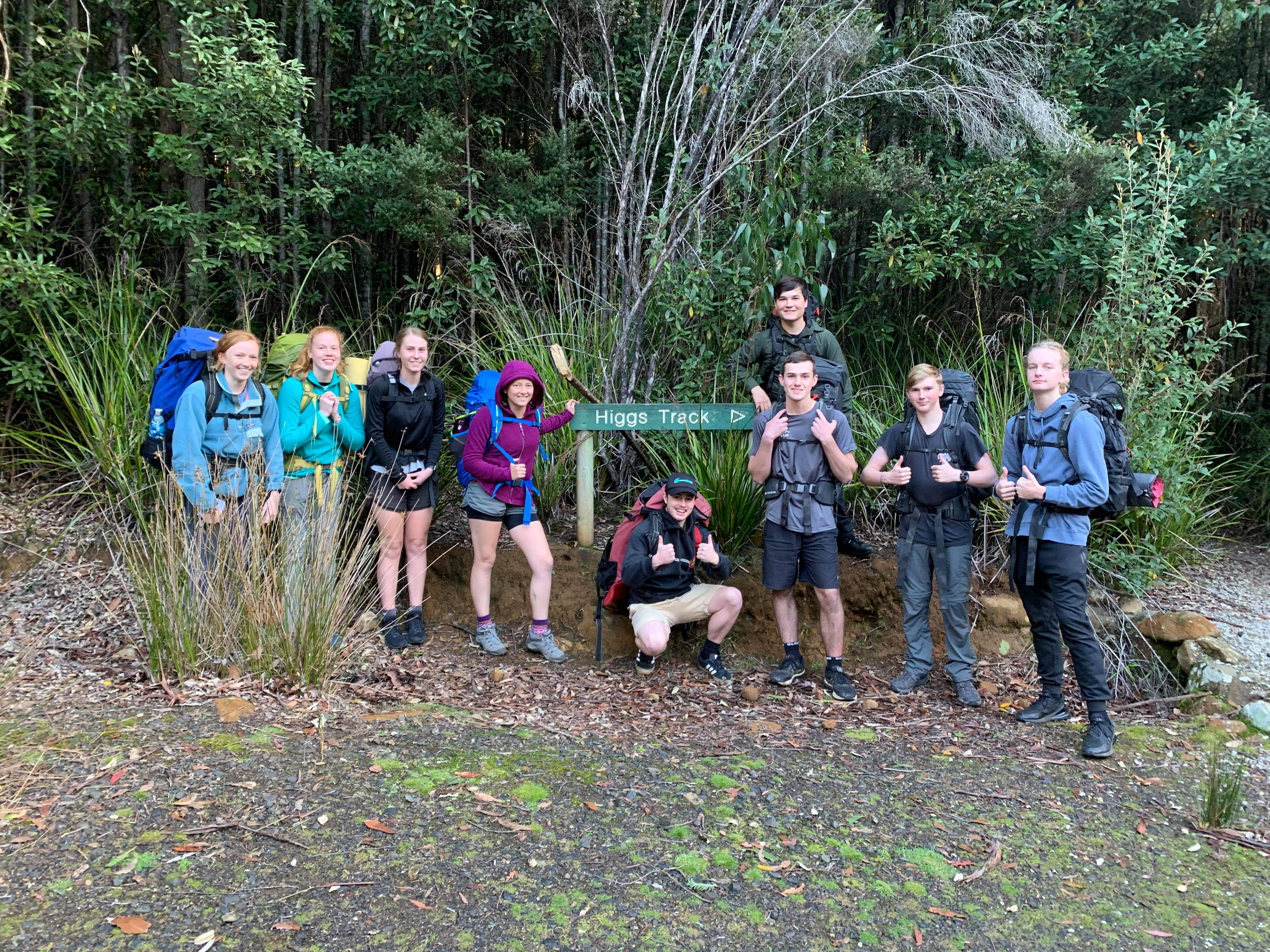 Group of eight young hikers with backpacks posing beside a green sign that reads 'Higgs Track' at a forest trailhead.