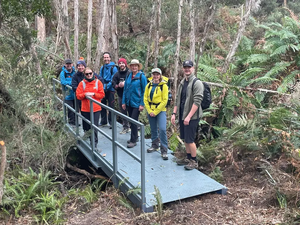 Group of eight hikers standing on a small metal bridge in a dense forest with ferns and tall trees.