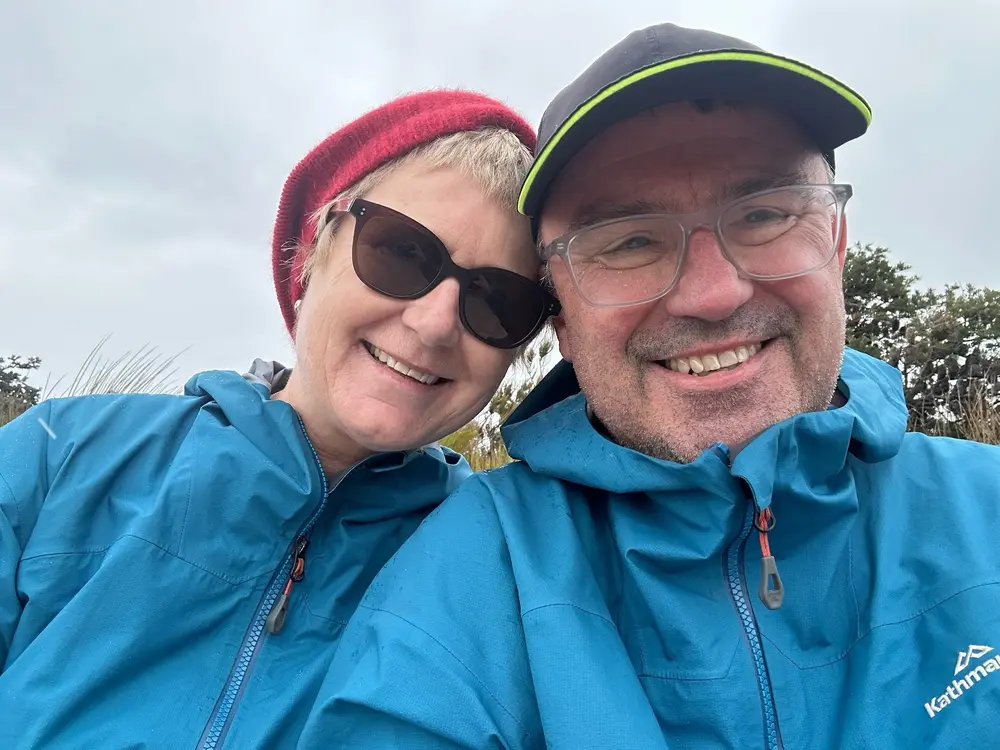 Smiling middle-aged couple wearing blue rain jackets outdoors, with the woman in sunglasses and a red beanie, and the man in clear glasses and a black cap.