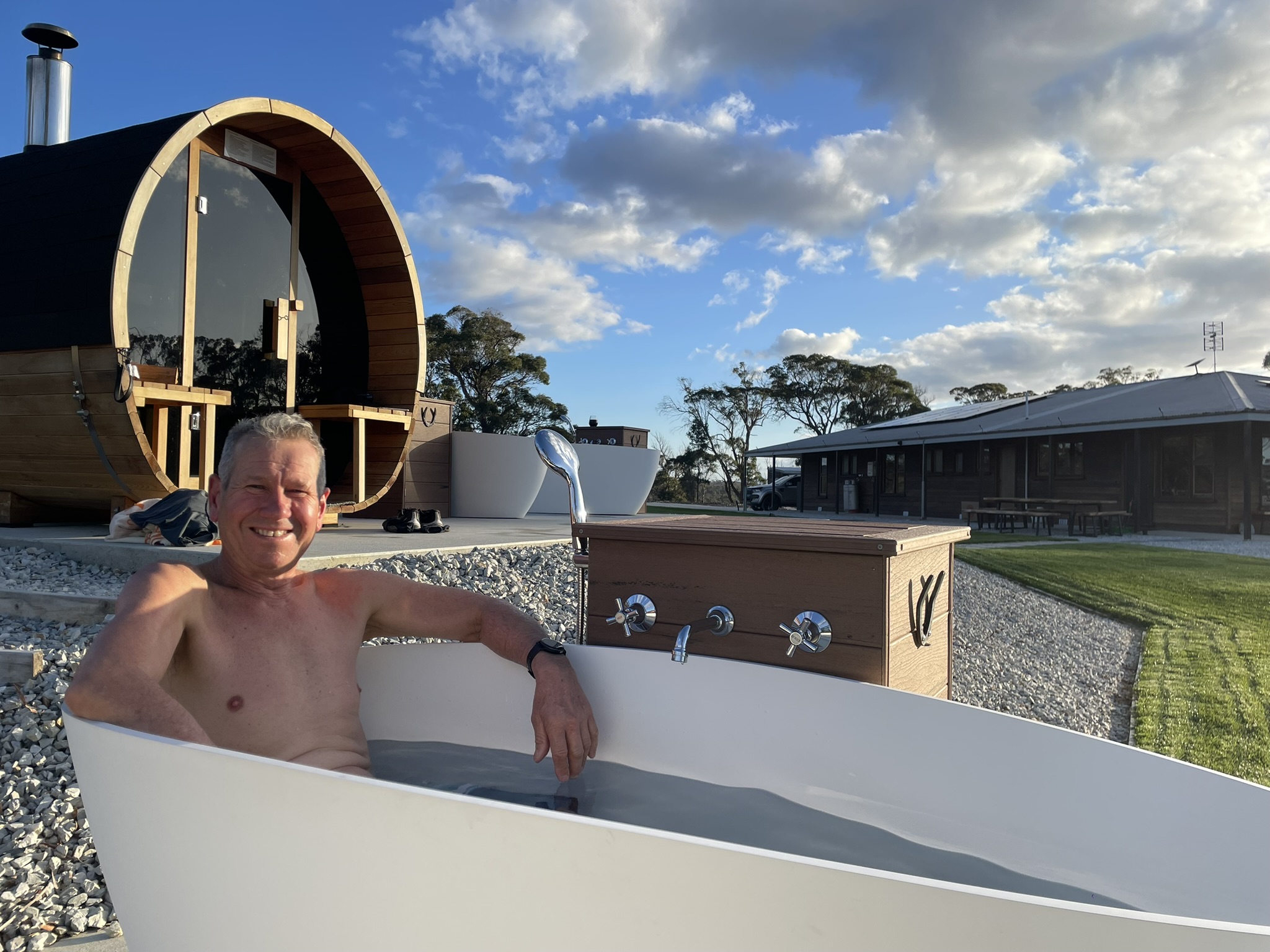 Smiling shirtless man relaxing in an outdoor white bathtub with a wooden barrel sauna and a building in the background under a partly cloudy blue sky.