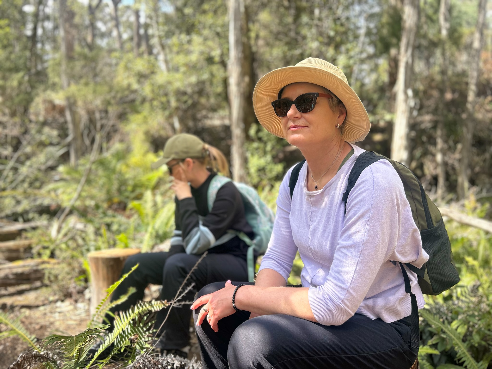 Two women resting in a forested area, one wearing a sun hat and sunglasses sitting on a log in the foreground, the other with a baseball cap and backpack blurred in the background.