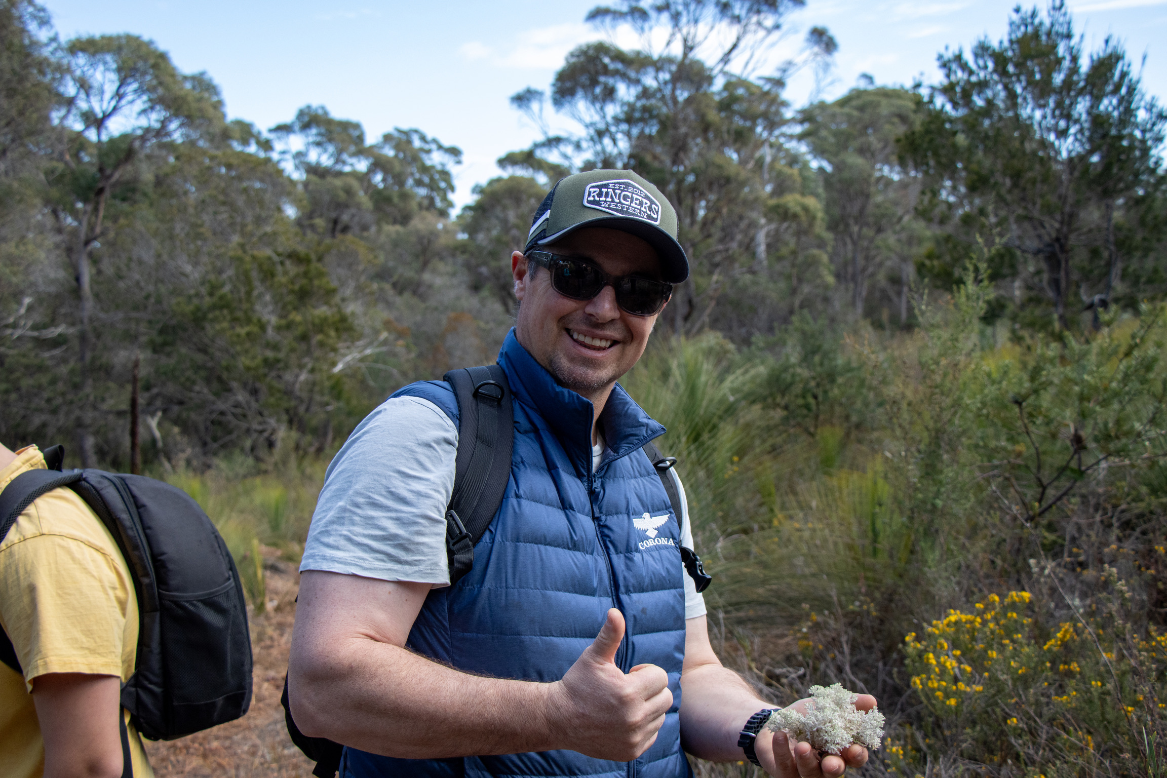 Man wearing sunglasses, a cap, and a blue vest hiking with a backpack, holding white lichen and giving a thumbs up in a forested area.
