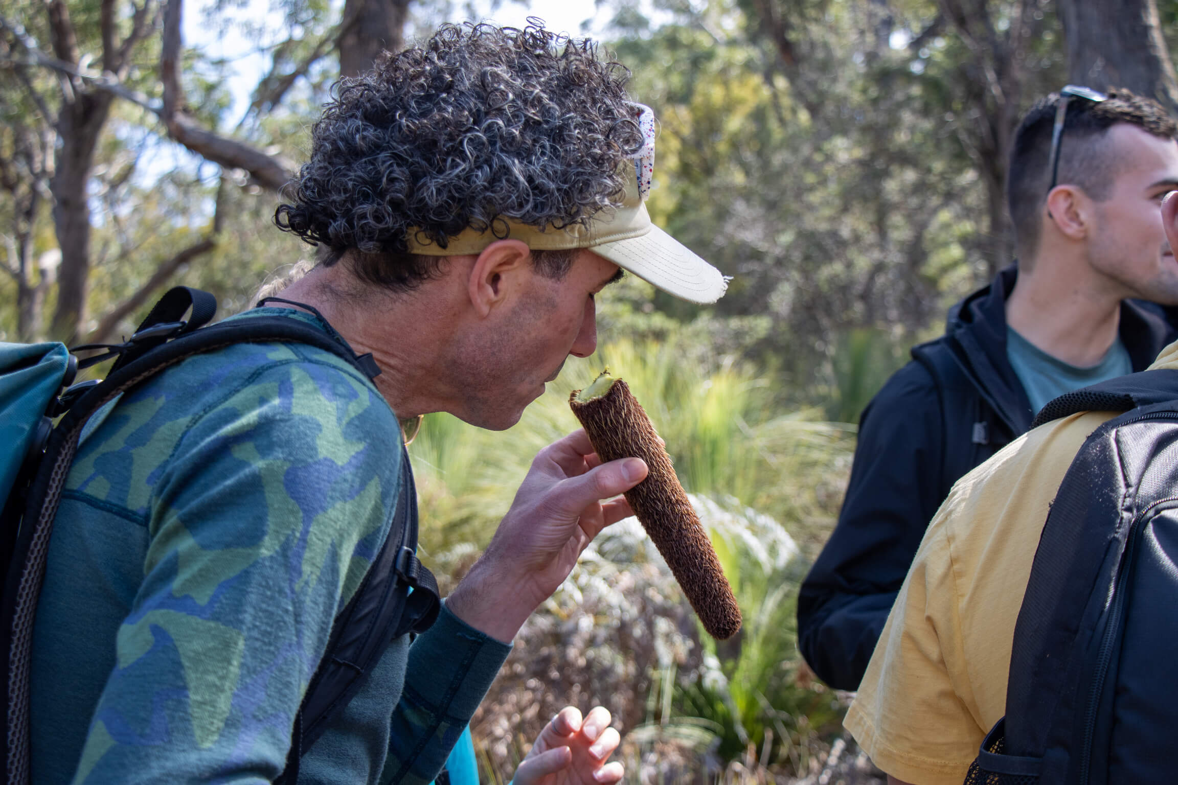 Man in a green camouflage shirt and beige cap holds and inspects a long brown seed pod in a forest setting with other people around.