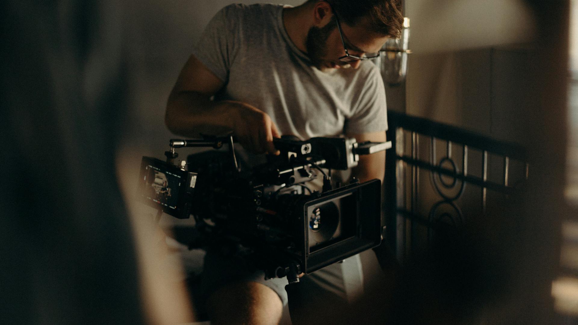 Man wearing glasses adjusts a professional video camera indoors in low light.